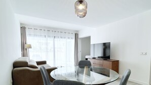 Ein modernes Wohnzimmer mit einem Glastisch, grauen Polsterstühlen, einer Couch, einem Fernseher auf einer Holzkommode und leichten Vorhängen am Fenster.