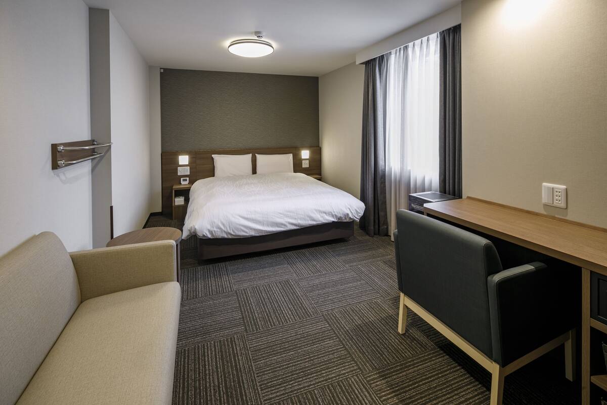 A hotel room with a bed, a desk, a chair, a sofa, and a carpeted floor.