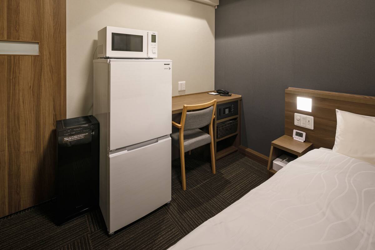 A hotel room with a bed, a desk with a chair, a microwave, a refrigerator, a trash bin, and a bedside table with a lamp.