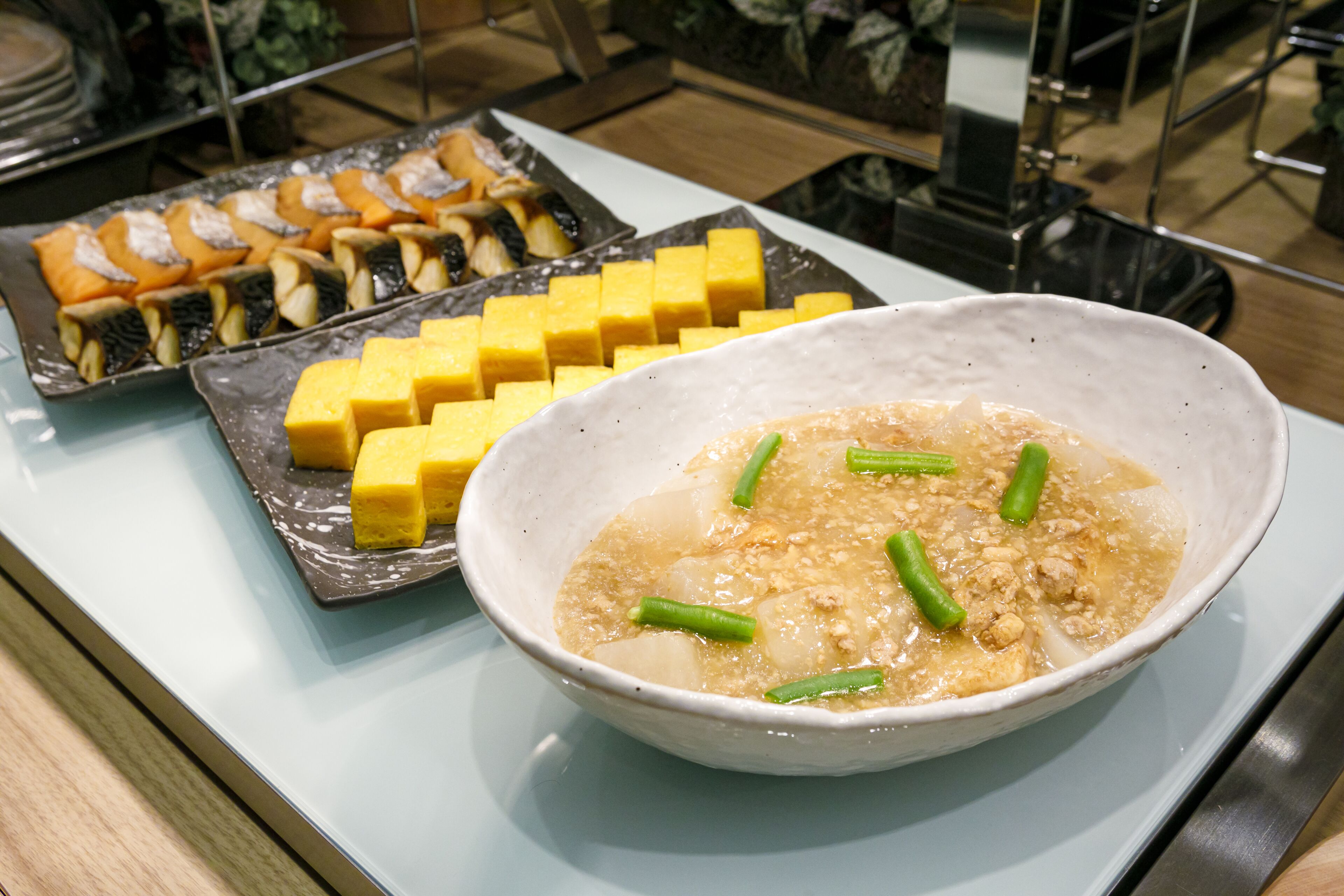 A buffet table with a variety of dishes including a bowl of soup with green vegetables and tofu, and several platters of yellow and orange food items.