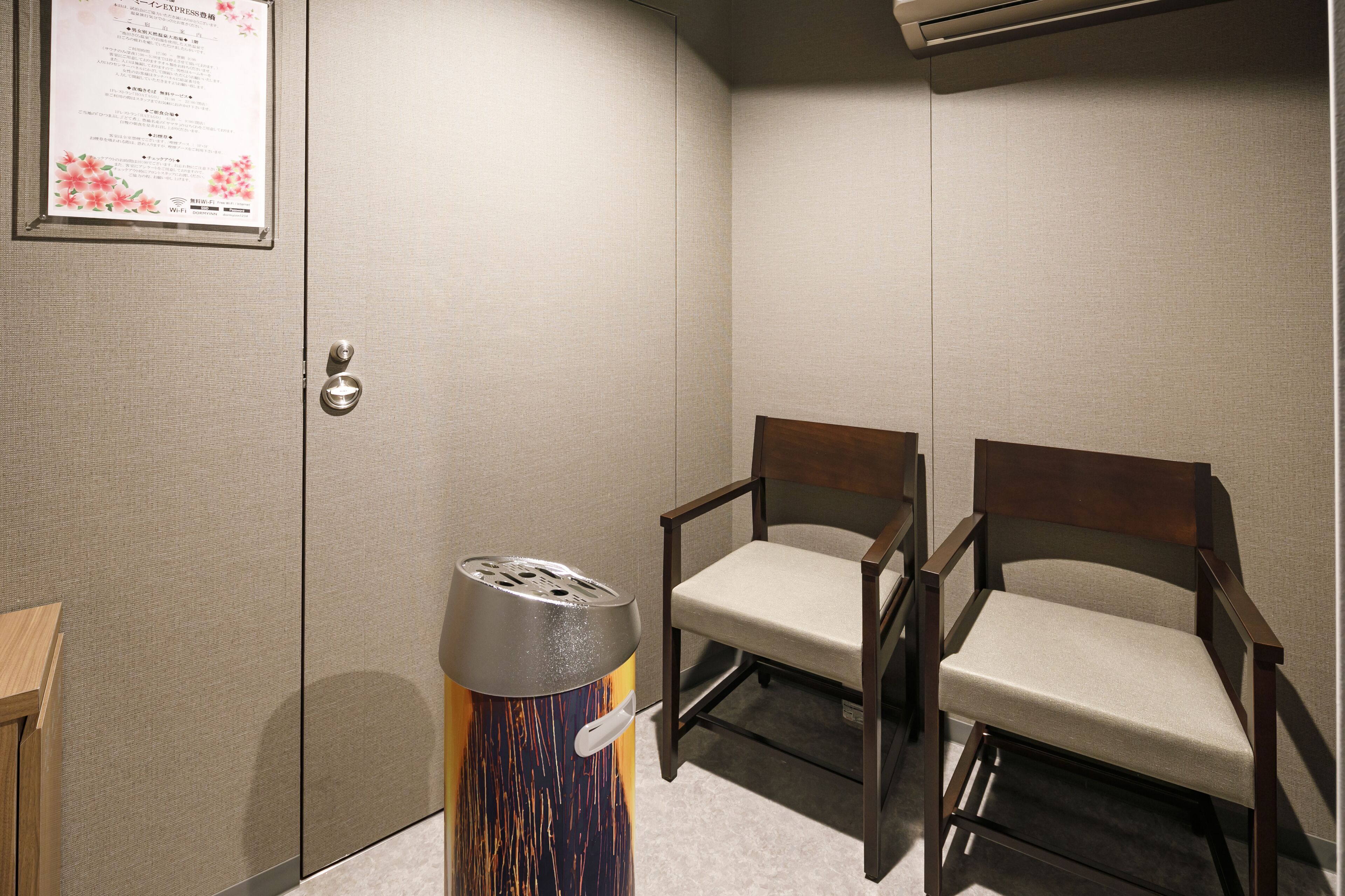 A small, private room with a wooden chair, a trash bin, and a wall-mounted air conditioning unit.