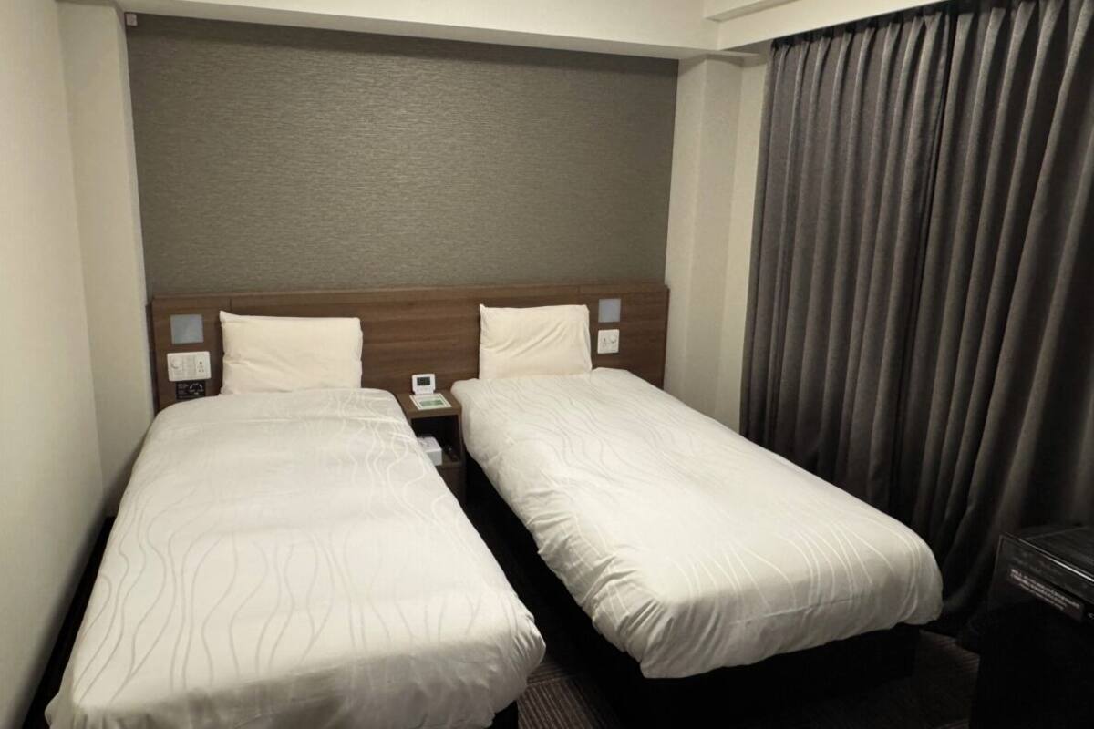 A hotel room with two single beds, a wooden headboard, a nightstand with a phone, and a large window with curtains.