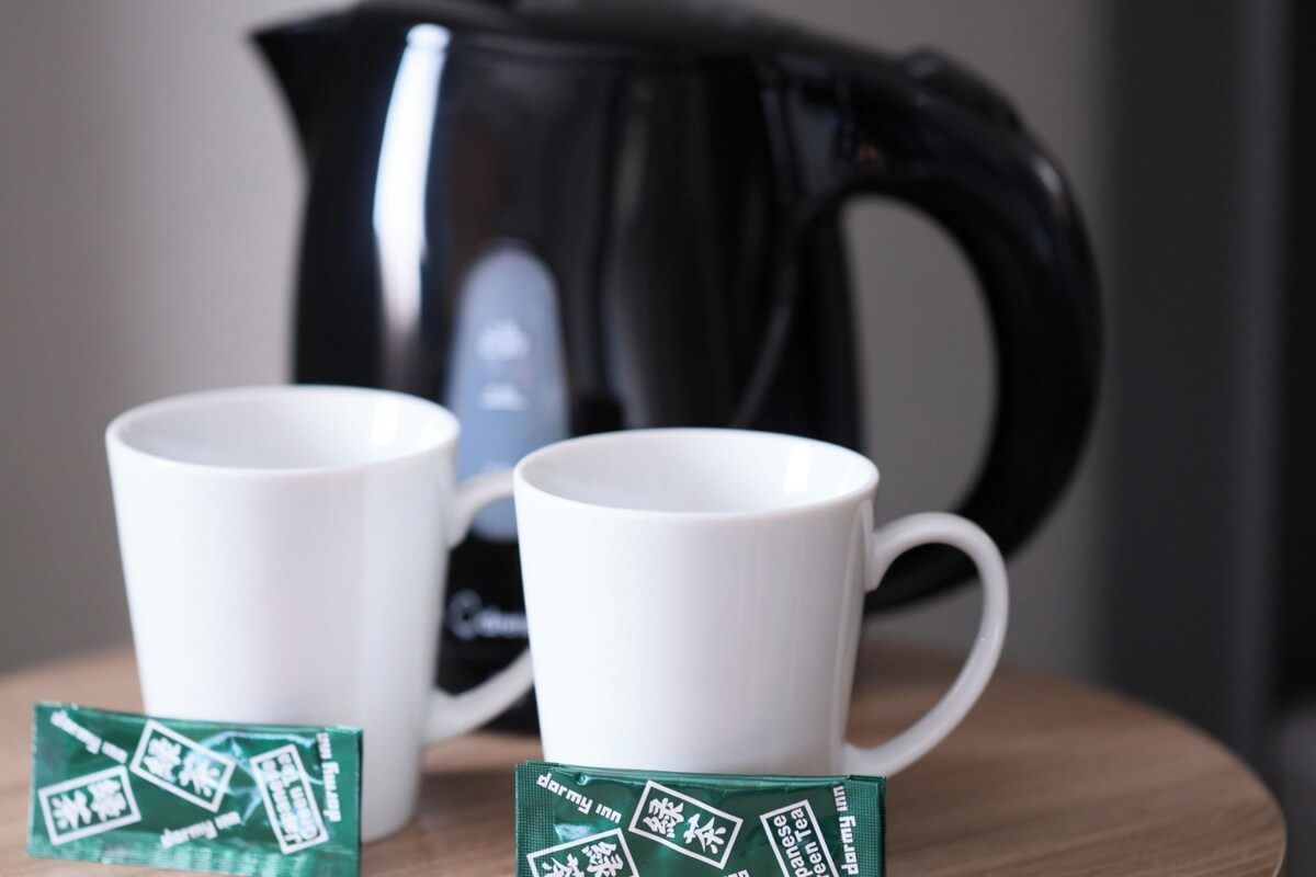 Two white mugs on a wooden surface with a green tea packet in front of each. A black kettle is in the background.