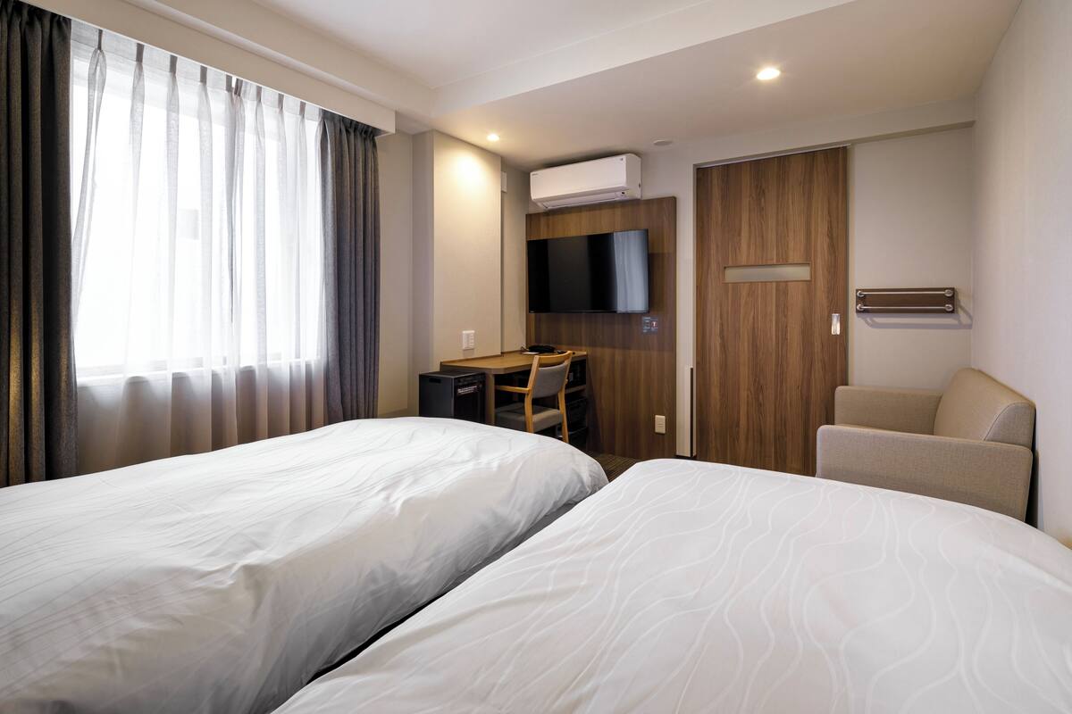 A hotel room with two beds, a desk, a chair, a TV, and an air conditioning unit.