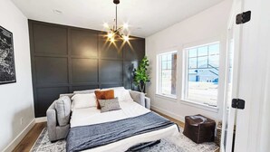 Une chambre moderne avec un grand lit, une tête de lit sombre, un luminaire suspendu et une vue sur l’extérieur par une fenêtre.