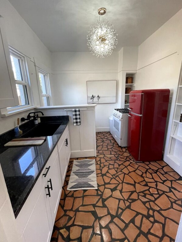 Una cocina con armarios blancos, encimera negra, refrigerador rojo, alfombra estampada y candelabro.