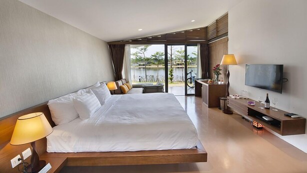 A modern hotel room with a large bed, a TV, a desk with a lamp, and a view of the outdoors. - Flamingo Dai Lai Resort Vila Tu Nhan BBQ (Vinh Phuc)