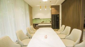 A conference room with a long table and eight chairs, a kitchen area in the background, and a wooden panel wall.