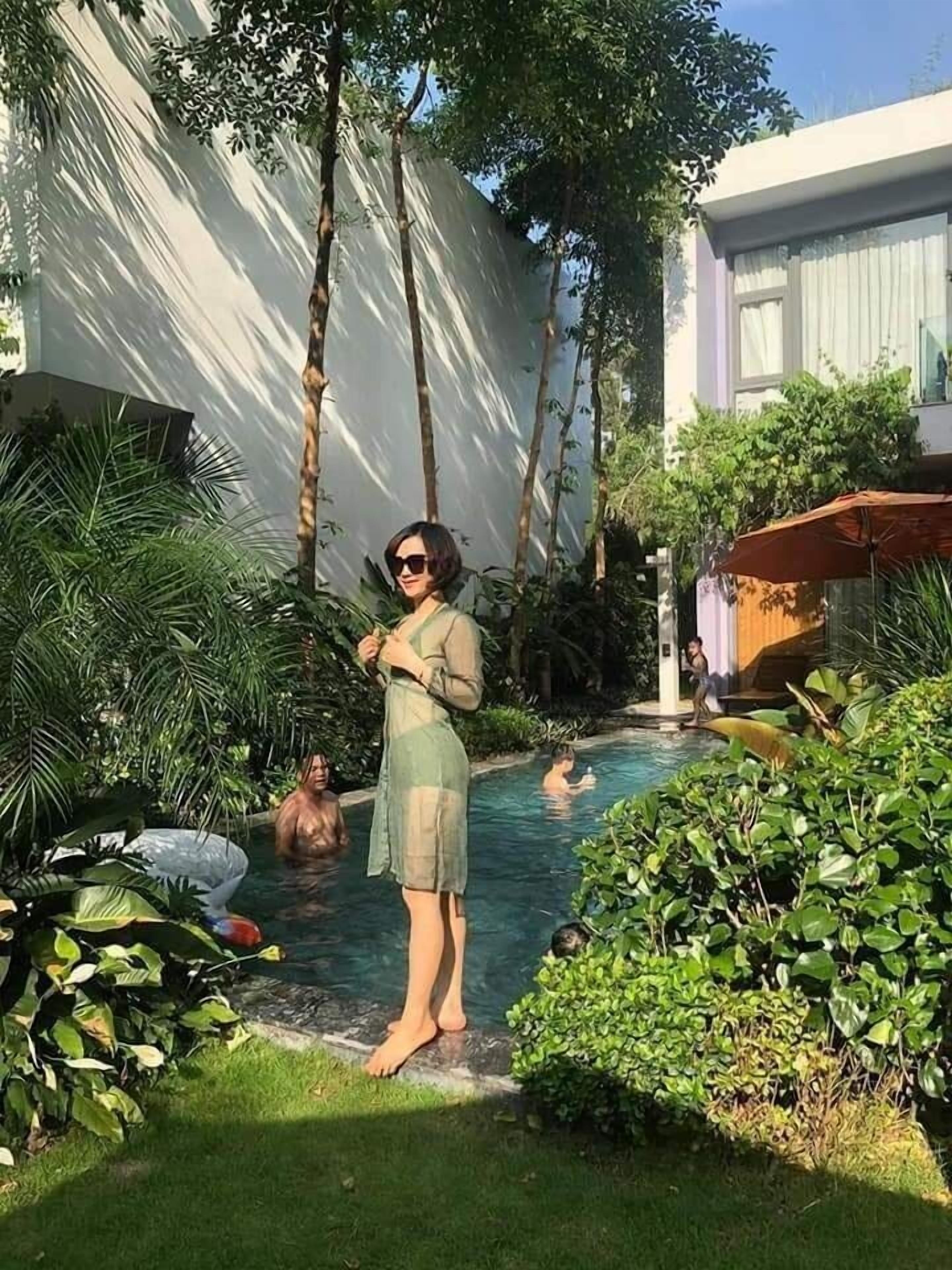 A pool area with a person standing by the pool, surrounded by lush greenery and a building in the background.