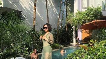 A pool area with a person standing by the pool, surrounded by lush greenery and a building in the background.