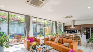 A modern living room with orange sofas, a coffee table with drinks, and a view of a kitchen and outdoor area with plants. - Flamingo Dai Lai Resort Vila Tu Nhan BBQ (Vinh Phuc)