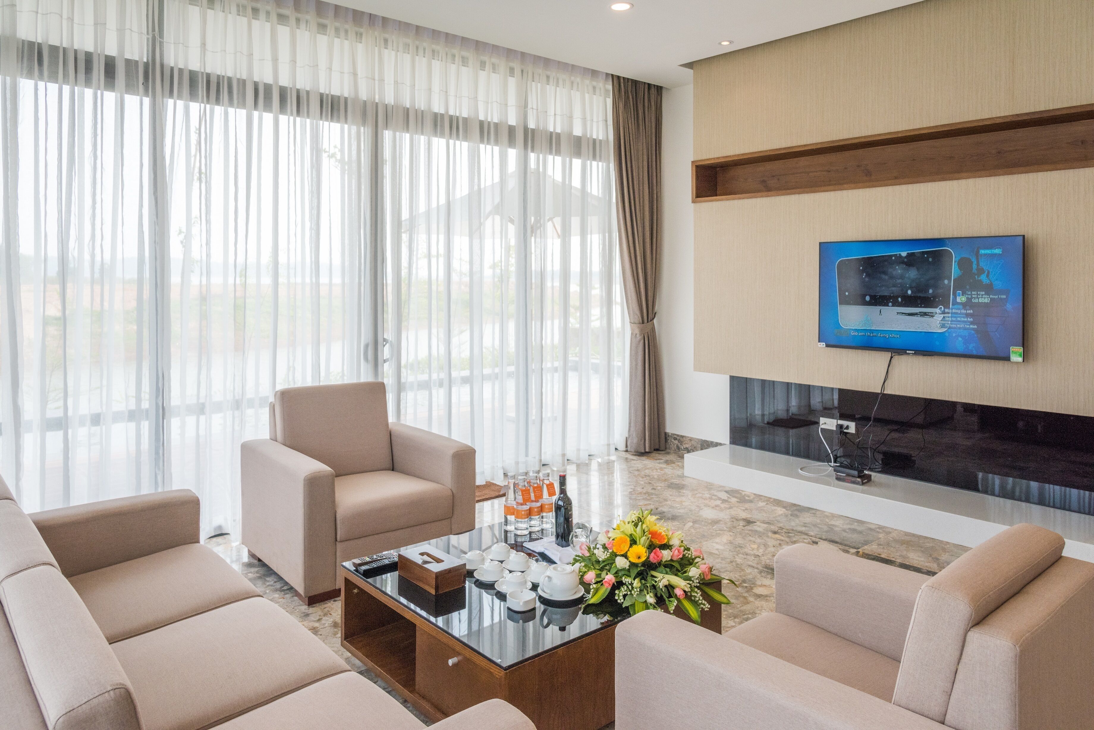 A modern living room with a sofa set, a coffee table with tea cups and a flower arrangement, and a flat-screen TV mounted on the wall.