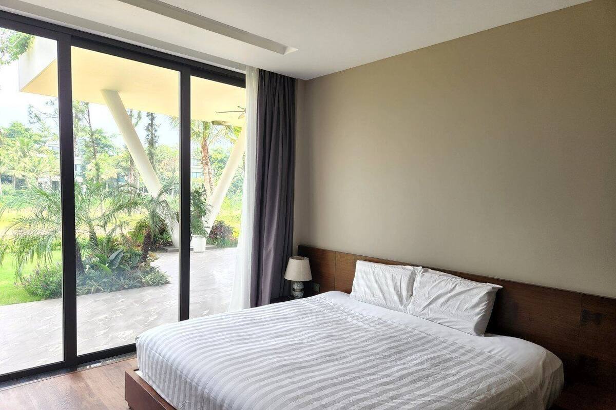 Une chambre d’hôtel avec un grand lit, une table de chevet, une lampe et une vue sur la verdure, visible à travers une porte-fenêtre coulissante.