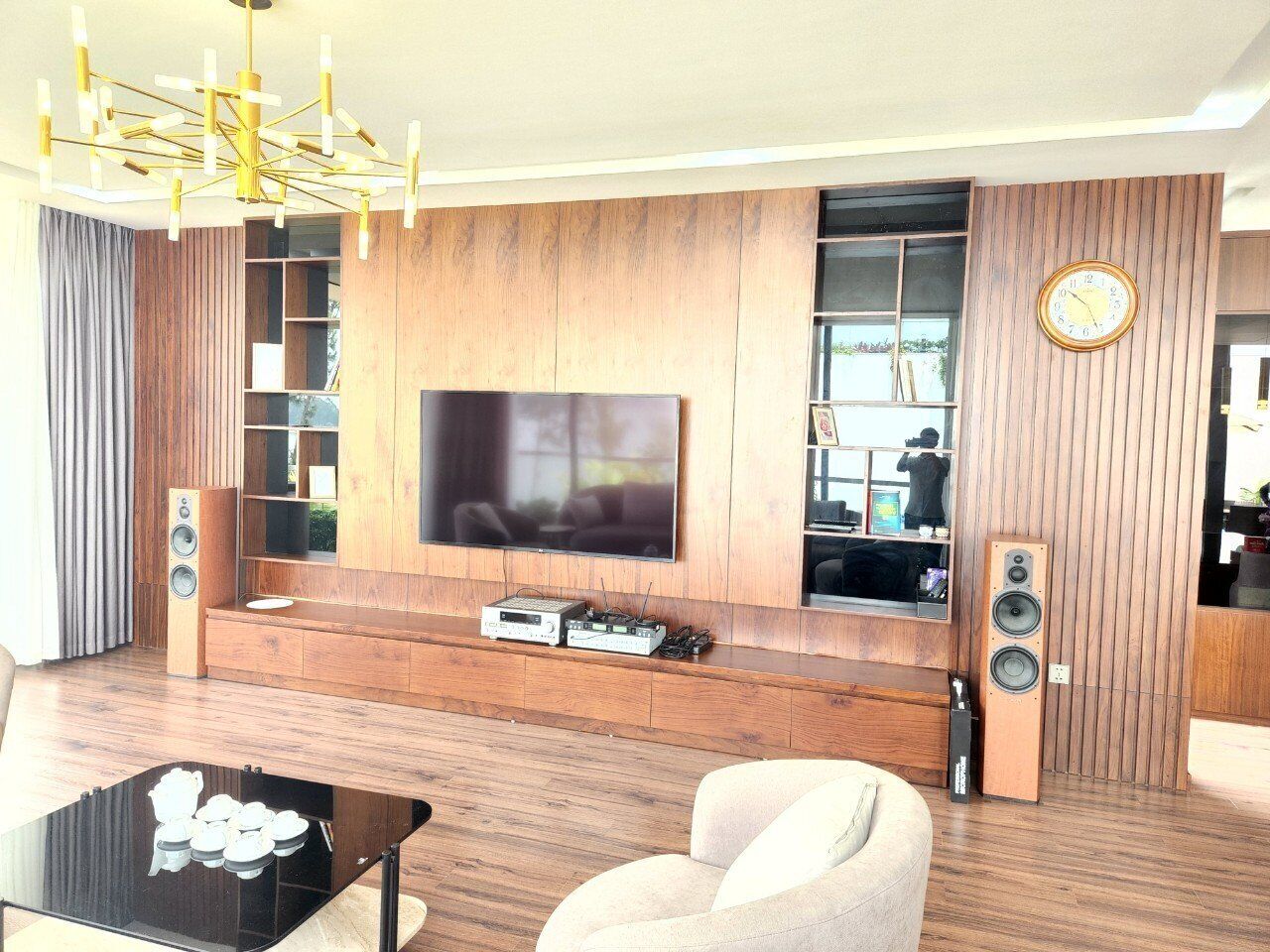Un salon moderne doté d’un grand téléviseur à écran plat, de murs recouverts de panneaux en bois, d’un système audio intégré et d’un lustre élégant.