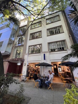 Un'area esterna con tavoli e sedie, un edificio con diverse finestre e un albero sullo sfondo.