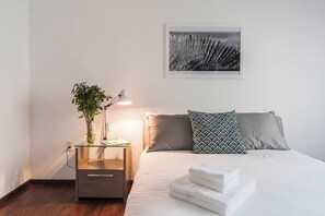 A neatly made bed with pillows, a bedside lamp, a small table with a plant, and a framed picture on the wall.