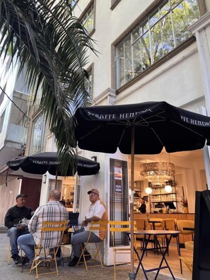 Zona esterna con tavoli e sedie sotto ombrelloni, una palma e l'ingresso di un edificio con un cartello.