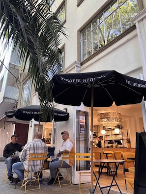 An outdoor seating area with tables and chairs under umbrellas, a palm tree, and a building entrance with a sign.
