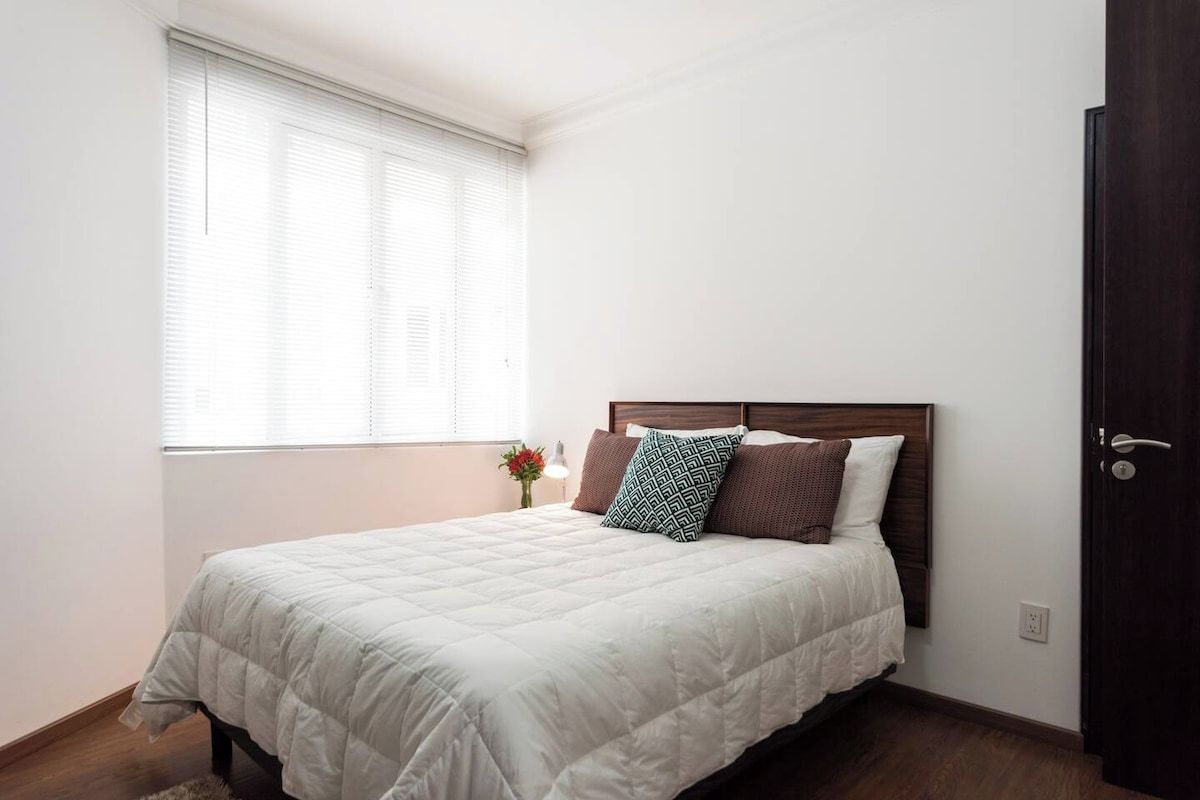 Una camera da letto con un letto, una finestra con persiane, una porta e un piccolo vaso con fiori.