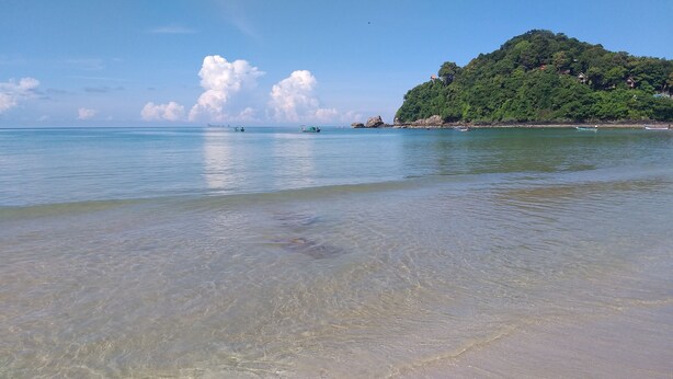 A beach with clear shallow waters, boats, and a forested hill in the background. - Cashewnut Tree Resort (Ko Lanta)