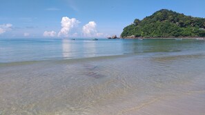 Uma praia com águas cristalinas e pouco profundas, barcos e uma colina arborizada ao fundo.