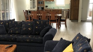 A modern living room with a dark blue sectional sofa, wooden dining table, and chairs. A kitchen with wooden cabinets and a microwave is visible in the background.