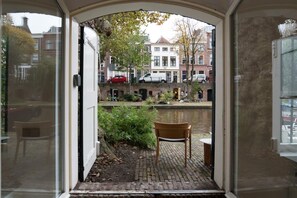 A view through a doorway to a canal-side area with a chair and a small table, buildings in the background, and a red car parked on the street.