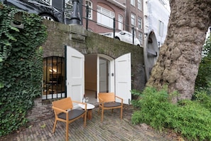 A cozy outdoor seating area with two wooden chairs and a small table, surrounded by a brick wall and greenery.
