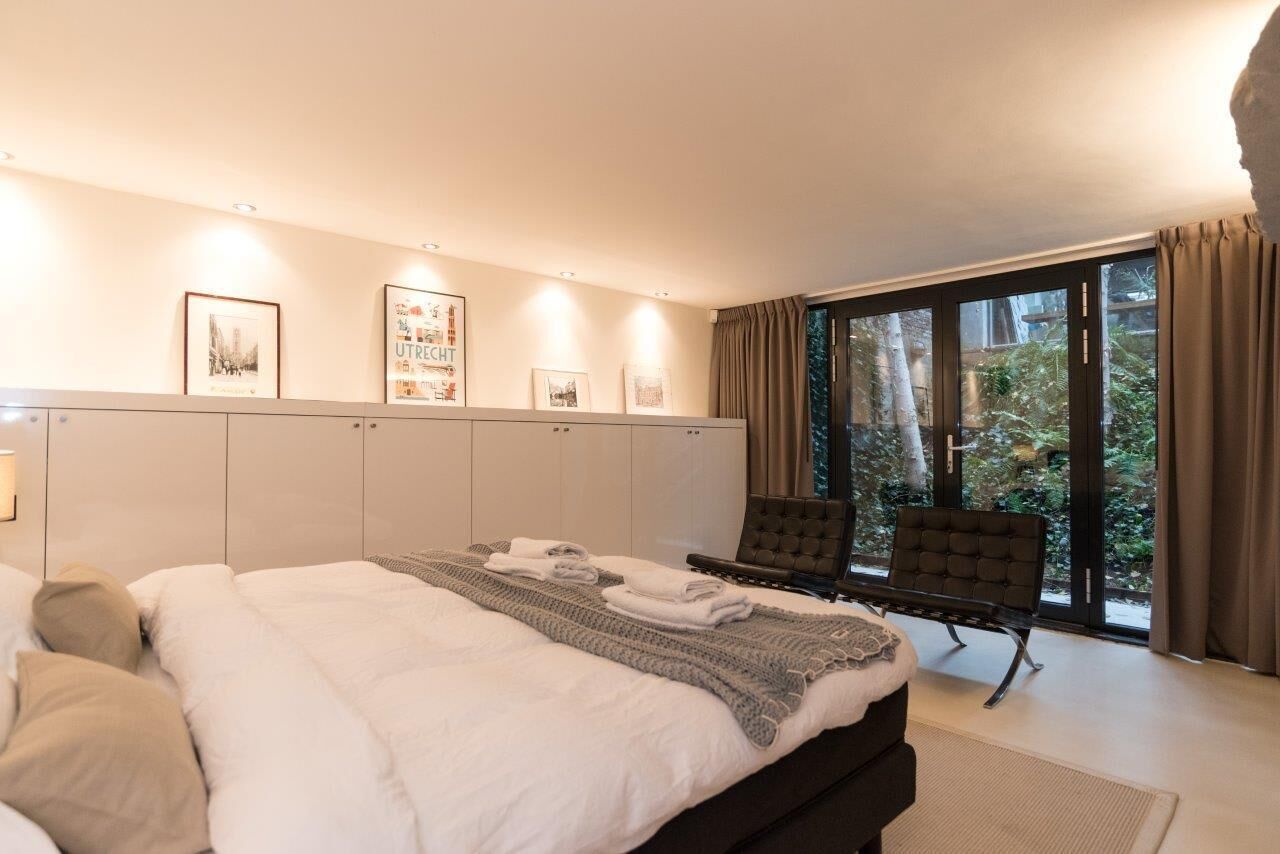 A bedroom with a large bed, a chair, a wardrobe, and a sliding glass door to a garden.
