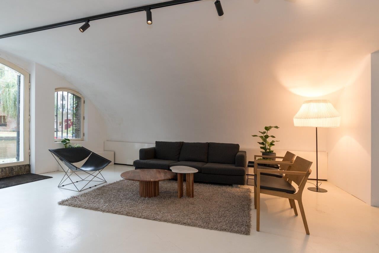 Ein modernes Wohnzimmer mit einer schwarzen Couch, einem Holztisch, einem kleinen Beistelltisch, einem Sessel, einer Stehlampe und einem großen Fenster mit Blick auf Grünflächen.