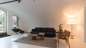 Ein modernes Wohnzimmer mit einer schwarzen Couch, einem Holztisch, einem kleinen Beistelltisch, einem Sessel, einer Stehlampe und einem großen Fenster mit Blick auf Grünflächen.