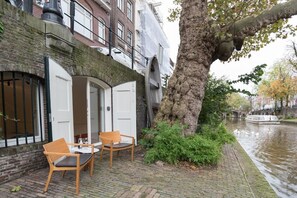 A wooden bench and table set up on a brick-paved area next to a canal, with a tree and a building in the background. - Canallofts - The Wharf Wellness (Utrecht)