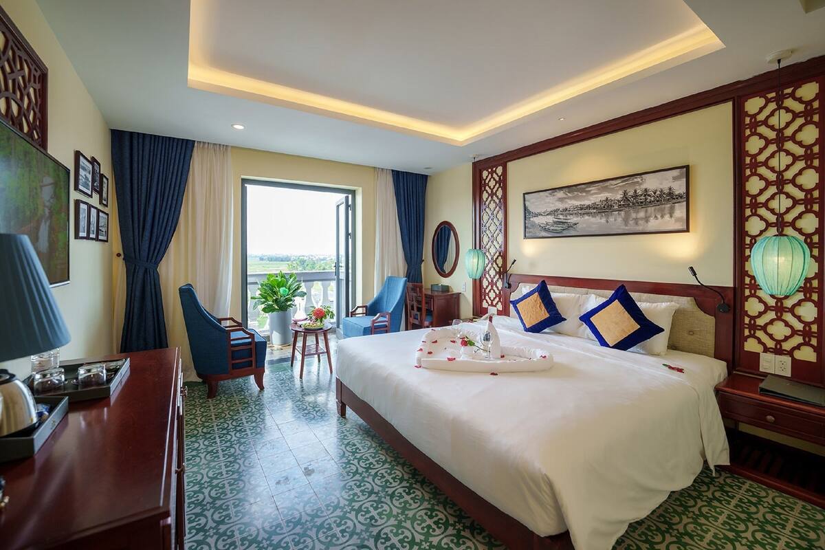Ein Hotelzimmer mit einem großen Bett, einem Schreibtisch, einem Sessel, einem kleinen Tisch mit Pflanze und einem Balkon mit Blick.