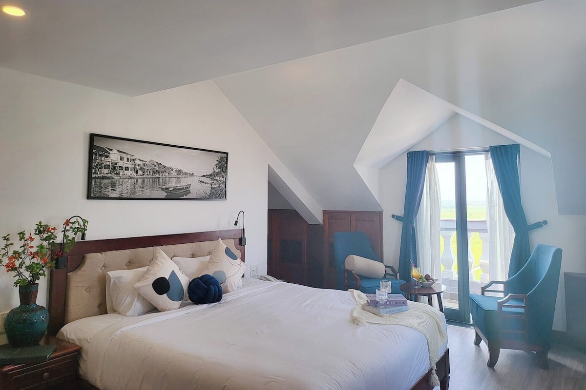 Ein Hotelzimmer mit einem großen Bett, einem Bild an der Wand, einer Vase mit Blumen und Blick ins Freie durch ein Fenster.