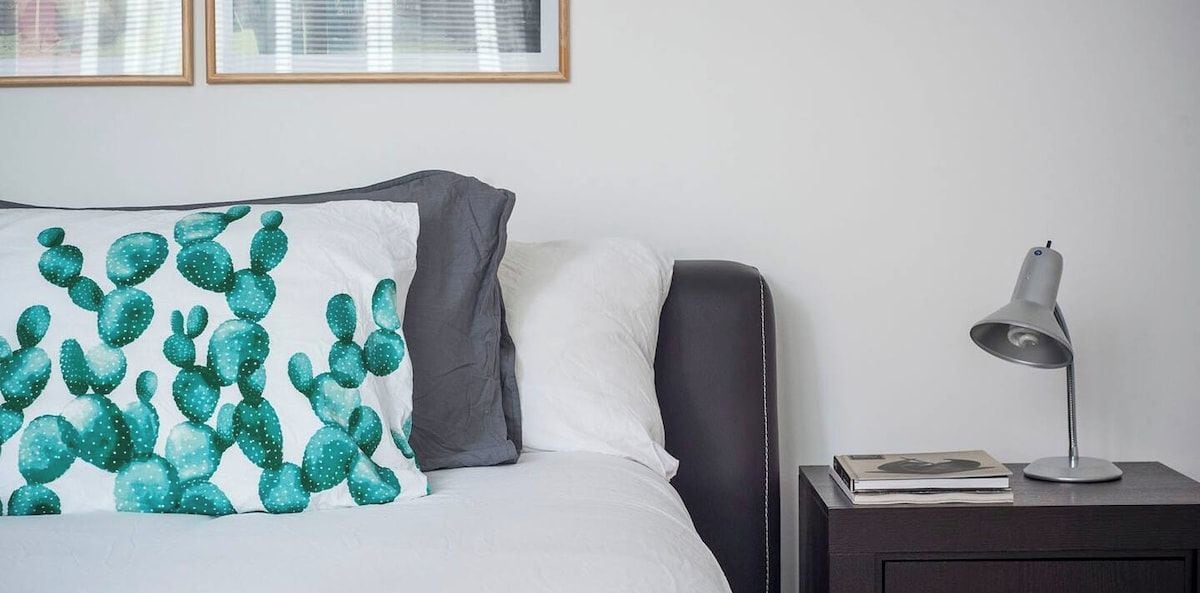 Un letto rifatto con cura, un cuscino a tema cactus, una lampada da comodino e un quadro appeso al muro.