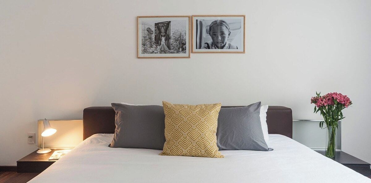 A neatly made bed with two pillows, a bedside lamp, and a vase with flowers on a nightstand.
