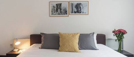 A neatly made bed with two pillows, a bedside lamp, and a vase with flowers on a nightstand.