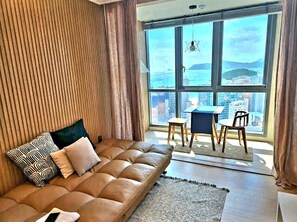 Sala moderna com sofá de couro, uma mesinha e vista para a cidade através de janelas amplas.