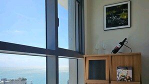 Une pièce dotée d’une grande fenêtre donnant sur la mer et les bâtiments. On y trouve un meuble en bois sur lequel sont posés une bouteille de vin et des verres.