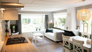 A modern kitchen with a gas stove, a white dining table with chairs, a sofa, and large windows with curtains.