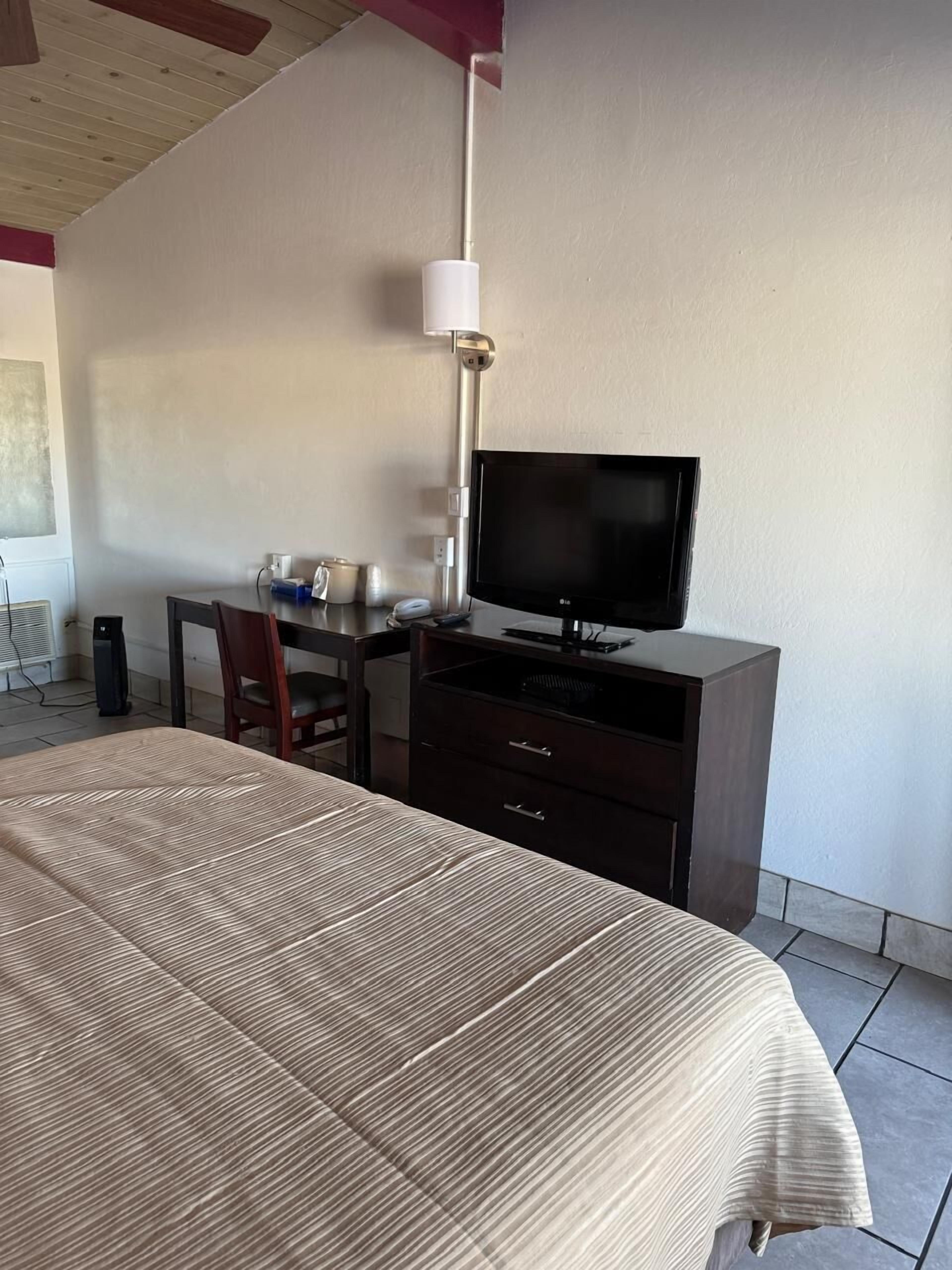 Quarto de hotel com uma cama, uma mesa de trabalho com televisão, uma cadeira e um abajur.