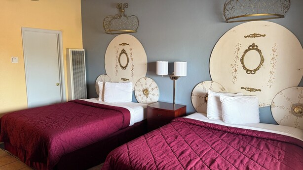 Habitación de hotel con dos camas, cada una con una colcha roja, una mesita de noche con dos lámparas y apliques decorativos en la pared.