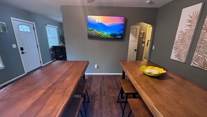 Un comedor con una larga mesa de madera y bancos, un televisor de pantalla plana montado en la pared, y una puerta que da a otra habitación.