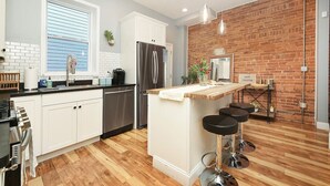 A modern kitchen with white cabinets, a stainless steel refrigerator, a wooden island with bar stools, and a brick accent wall.