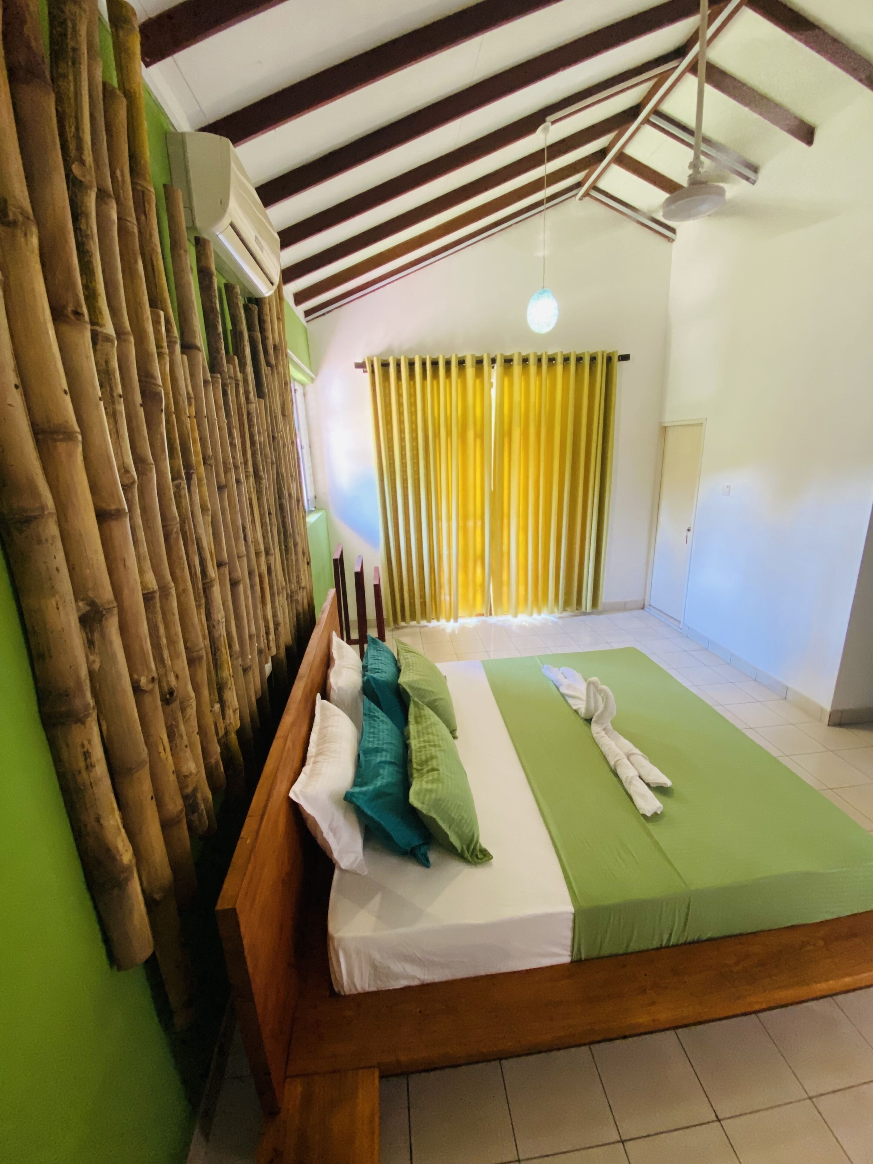 A room with a bed, a bamboo wall, a ceiling fan, and a window with yellow curtains.