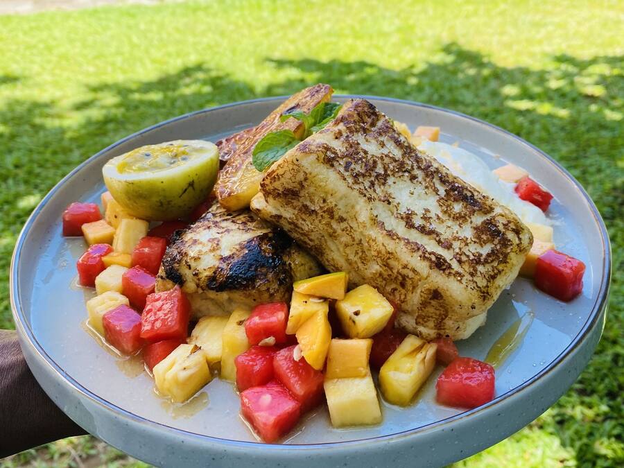 Un plato de pescado a la parrilla con trozos de mango y piña, sobre un área cubierta de hierba.