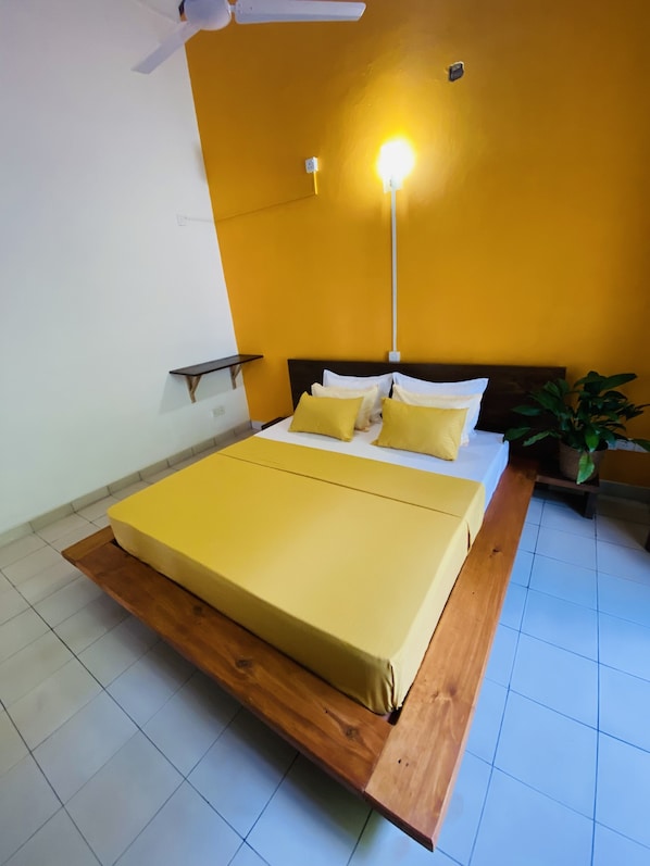 A bed with a wooden frame and a yellow mattress, a bedside table, a nightstand, a potted plant, and a ceiling fan. - SURF N SPICE (Dikwella)