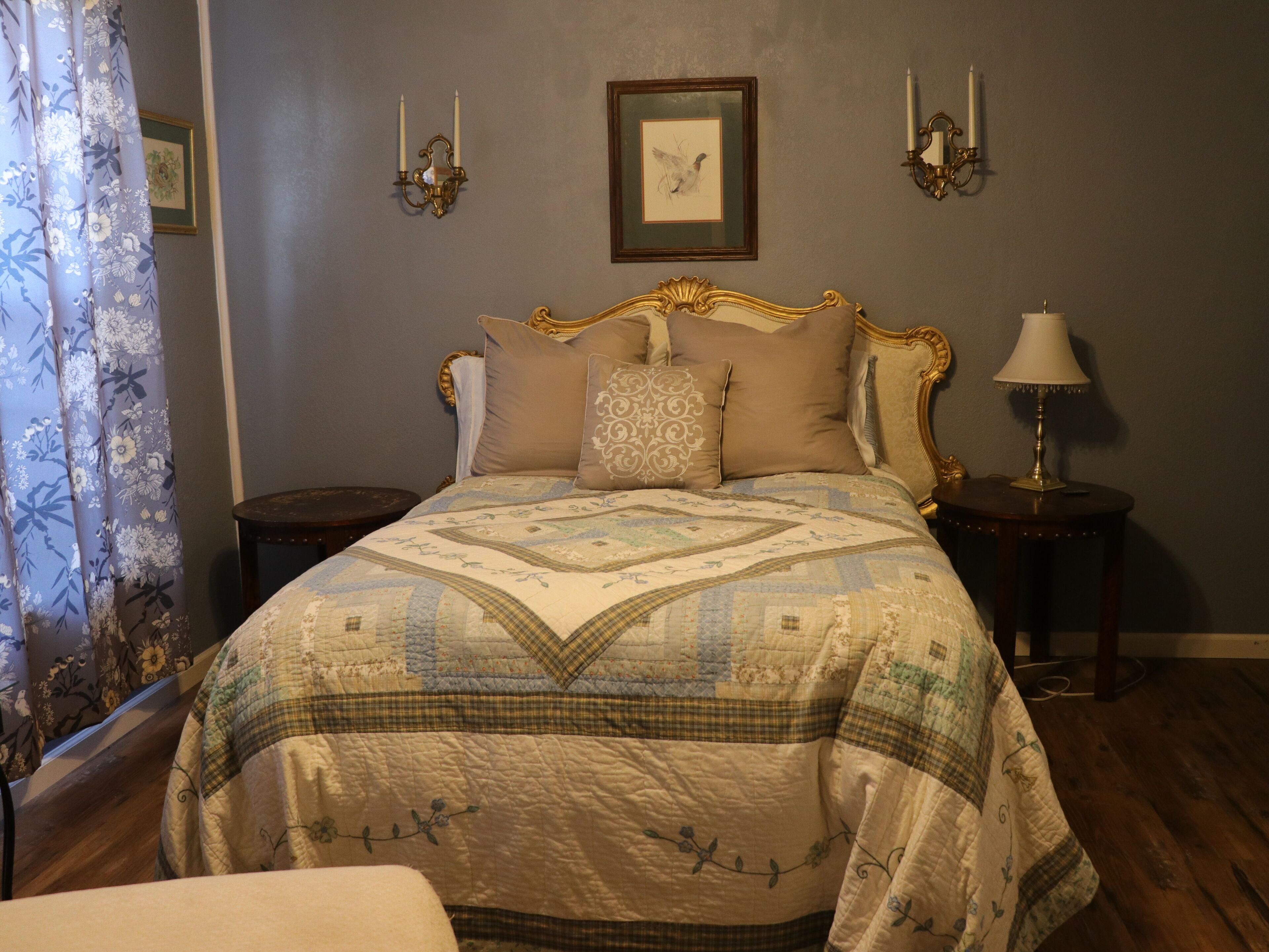 Une chambre à coucher avec un lit, une table de chevet, une lampe, un tableau encadré au mur et une fenêtre avec des rideaux.