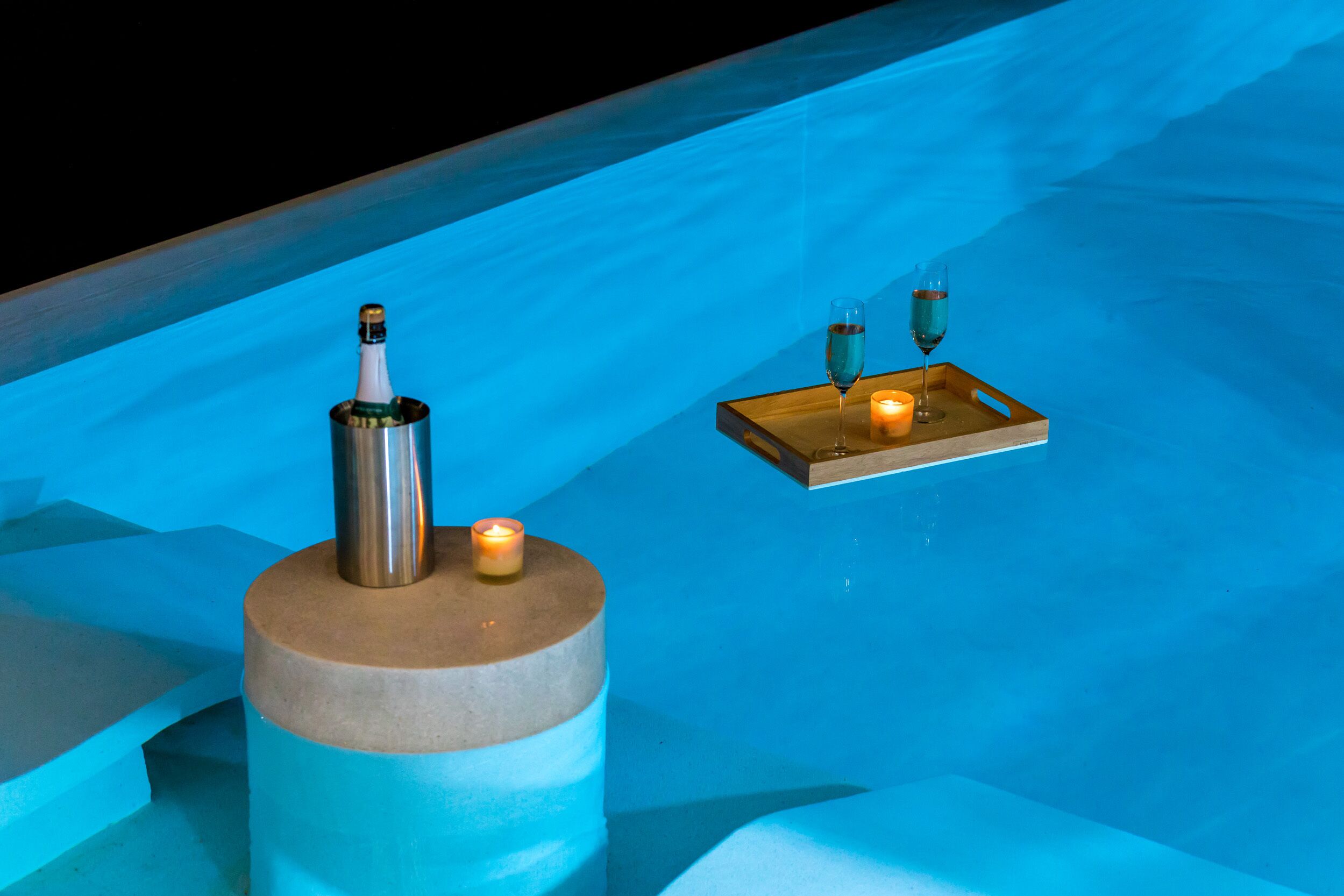 A blue-lit pool area with a champagne bottle, candle, and a tray with glasses.