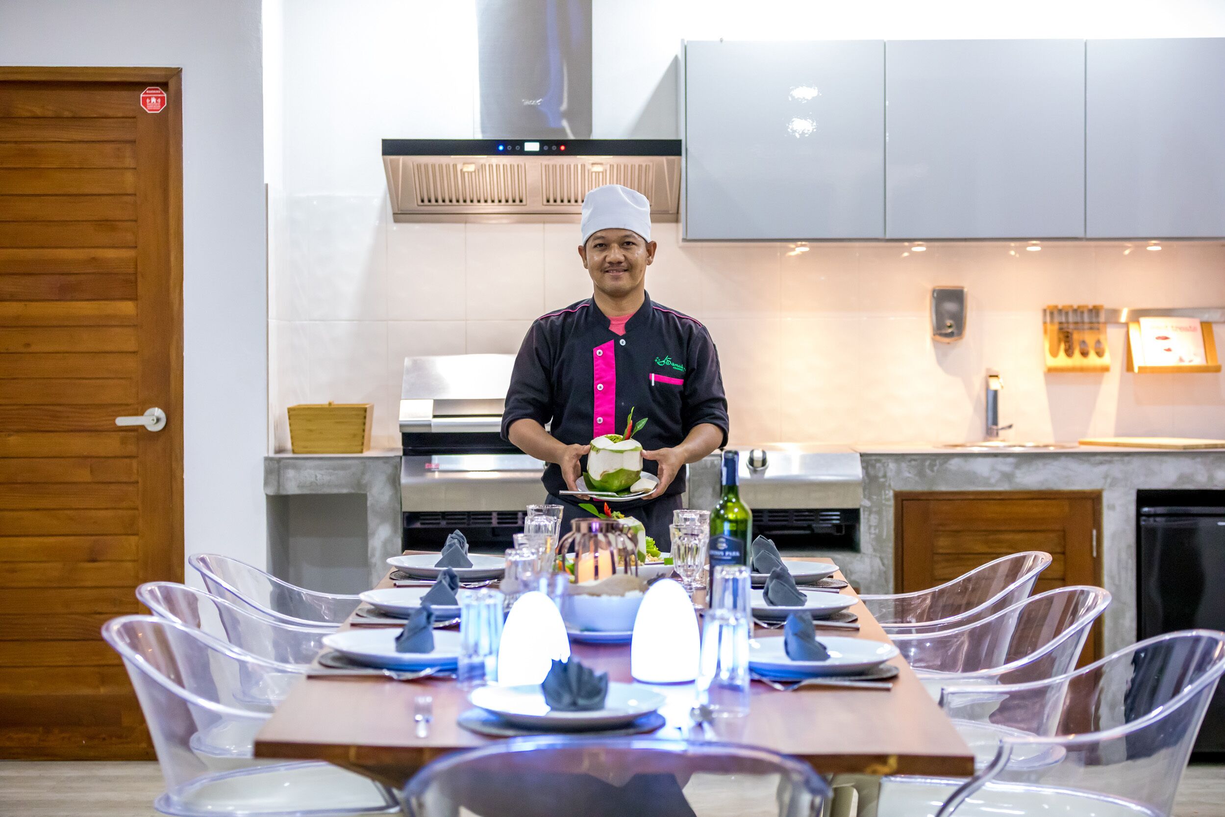 A chef in a kitchen holding a cake, with a dining table set with clear chairs and plates, and a modern kitchen in the background.